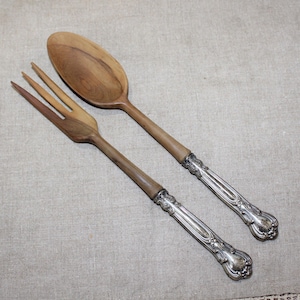 May include: A salad serving set consisting of a wooden spoon and a wooden fork. Each utensil features a polished brown wooden bowl and tines attached to an ornate, embossed silver-plated metal handle with intricate decorative detailing.