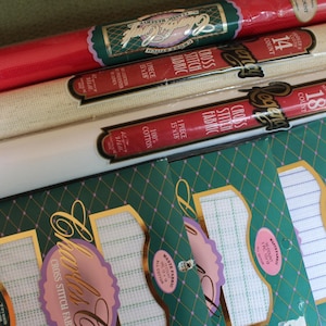 May include: Assortment of cross-stitch fabric rolls and packages. The rolls are red, cream, and white, with labels indicating 14 and 18 count fabric. Packages feature green diamond patterns and labels with the words "Charles" and "Cross Stitch Fabric".