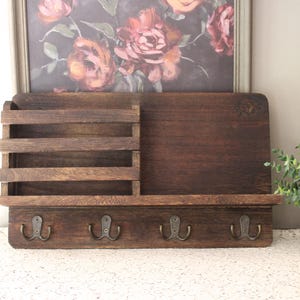 May include: A rustic wooden wall organizer with a dark brown finish. It features a mail sorter, a small shelf, and four metal hooks. The organizer is designed for entryway or home office use.