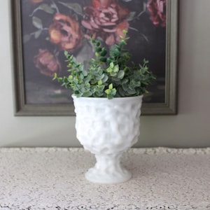 Könnte beinhalten: Eine weiße Milchglasvase auf einem Fuß mit einer strukturierten, unebenen Oberfläche. Die Vase ist mit einer kleinen, blättrigen grünen Kunstpflanze gefüllt, die aus mehreren Stielen und kleinen abgerundeten Blättern besteht und einen Kontrast bildet.