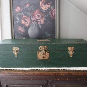 May include: A rectangular wooden storage box painted in a dark forest green finish. The box features a hinged lid with a central brass-toned latch and two smaller brass-toned side latches. A small brass-toned handle is mounted on the top lid.