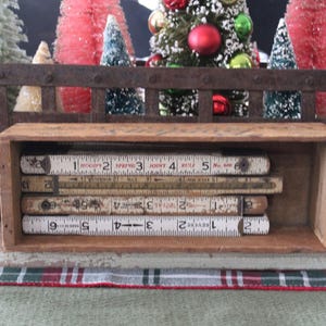 May include: A wooden box filled with vintage folding rulers, each marked with inch measurements. The rulers are stacked inside the box, with the numbers and markings clearly visible. The background features Christmas decorations.