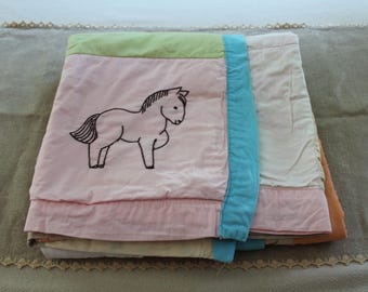 Puede incluir: Una colcha de retazos plegada que presenta un cuadrado rosa claro con el contorno de un caballo bordado en negro. La colcha incluye paneles de tela verde claro, azul brillante, crema y naranja, mostrando un diseño multicolor con costuras.