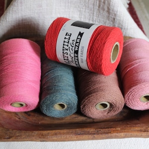 Könnte beinhalten: Fünf Garnrollen Maysville Fast Color Carpet Warp in verschiedenen Farben, darunter leuchtendes Rot, Pink, Altrosa und gedämpftes Blau. Jede Rolle hat einen Pappkern und ein weißes Papieretikett mit dem schwarzen Text 600 YARDS.