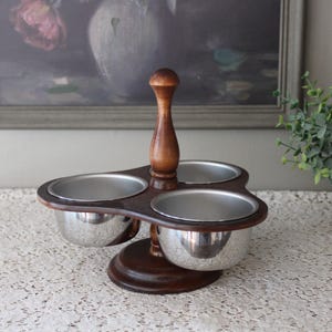 May include: A wooden condiment server with three stainless steel bowls. The server has a dark brown base and a handle. The bowls are arranged in a cloverleaf pattern. The server is designed for serving condiments.