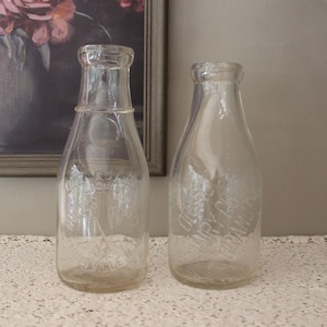 Puede incluir: Dos botellas de leche de vidrio transparente con texto en relieve. Las botellas tienen una forma clásica con un cuello estrecho y una base más ancha. Las botellas están vacías y parecen ser vintage. El fondo incluye una pintura y un mantel de encaje.