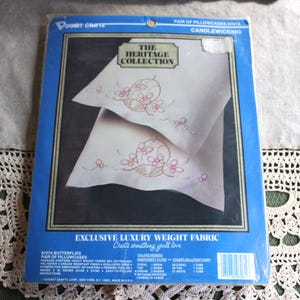 Könnte beinhalten: Eine blaue Verpackung mit einem Paar Kissenbezügen mit Schmetterlingsdesign. Die Verpackung trägt die Aufschrift "The Heritage Collection" und "Exclusive Luxury Weight Fabric". Die Kissenbezüge sind weiß mit rosa Stickereien.