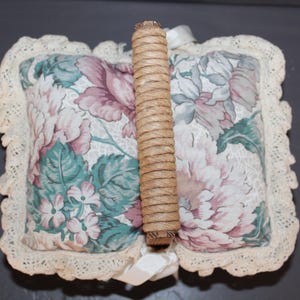 May include: A white floral fabric pillow with a woven wood handle. The pillow has a lace trim and a floral pattern in shades of pink, green, and blue.