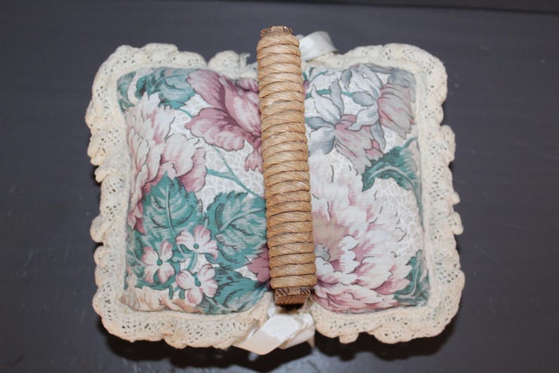 May include: A white floral fabric pillow with a woven wood handle. The pillow has a lace trim and a floral pattern in shades of pink, green, and blue.