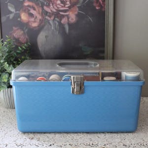 May include: A light blue sewing box with a clear lid, handle, and metal clasp. The box contains spools of thread and other sewing supplies. The box is sitting on a white lace tablecloth.