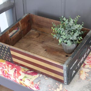 May include: A wooden crate with a painted American flag design on one side. The crate has metal reinforced edges and handles. A small potted plant sits inside the crate. The side of the crate reads "A-TREAT".