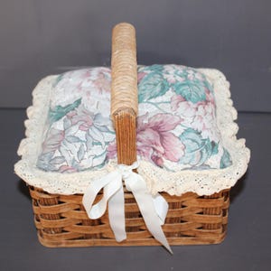 May include: A wicker basket with a floral fabric lining and a white lace trim. The basket has a wooden handle and a white ribbon tied around it.