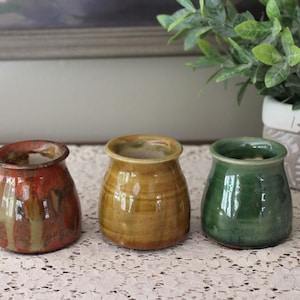 Op de afbeelding: Drie kleine, handgemaakte keramische potten in tinten rood, geel en groen. De potten hebben een afgeronde vorm met een licht uitlopende rand. Ze worden tentoongesteld op een kanten tafelkleed, met een plant op de achtergrond.