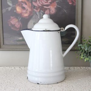 May include: A white enamelware coffee pot with a lid and a curved handle. The pot has a dark blue trim around the spout and lid. The pot is sitting on a white lace tablecloth. A framed floral painting is in the background.