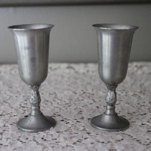 Op de afbeelding: Twee tinnen kelken met een uitlopende rand en een sierlijke steel. De kelken hebben een klassiek ontwerp en worden gepresenteerd op een wit kanten tafelkleed. De kelken zijn gemaakt van zilverkleurig metaal.