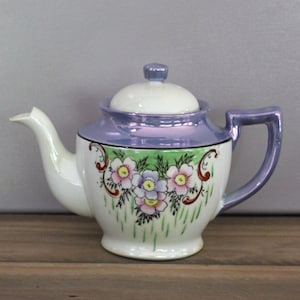 Puede incluir: Una tetera de porcelana vintage con cuerpo blanco, borde y asa iridiscentes lavanda y tapa a juego. La tetera está decorada con un diseño floral con flores rosas, moradas y amarillas sobre un fondo verde.