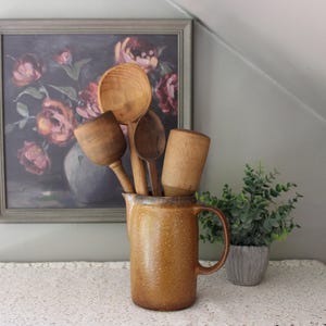 May include: A tall, cylindrical, mustard-yellow ceramic pitcher with a speckled texture and a side handle, containing a collection of various wooden kitchen utensils, including a large round spoon, a smaller spoon, and a wooden pestle.
