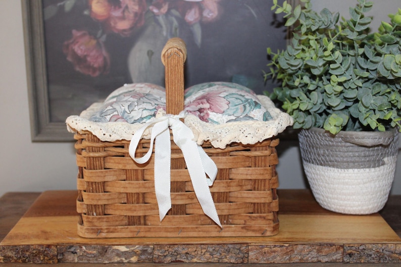 May include: A wooden basket with a floral fabric lining and a white ribbon. The basket has a handle and is sitting on a wooden surface.