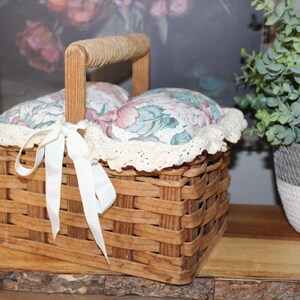 May include: A brown wicker basket with a floral fabric lining and a white lace trim. The basket has a wooden handle and a white satin ribbon tied around it.