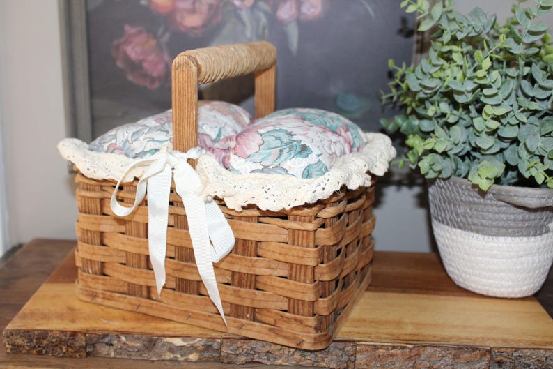 May include: A brown wicker basket with a floral fabric lining and a white lace trim. The basket has a wooden handle and a white satin ribbon tied around it.