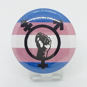 May include: A round button with a transgender pride flag design. The flag is pink, blue, and white with a black fist raised in the center of a transgender symbol.