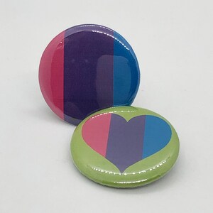 Könnte beinhalten: Zwei Anstecknadeln mit einem Thema der bisexuellen Stolz. Eine Nadel ist grün mit einem herzförmigen Design in den Farben der bisexuellen Pride-Flagge. Die andere Nadel ist lila, pink und blau mit vertikalen Streifen.