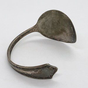 May include: A silver spoon bent into a bracelet shape. The spoon has a rounded bowl and a decorative handle.