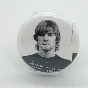 Puede incluir: Botón en blanco y negro con una foto de carnet de una persona con el pelo largo. El texto "AMERICAN" y "POLICE DEPT" es visible.