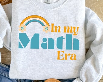 Può includere: Felpa grigio chiaro con la scritta "In my Math Era" in turchese e arancione. Un arcobaleno con simboli pi è sopra la parola "Math". La felpa è a girocollo.
