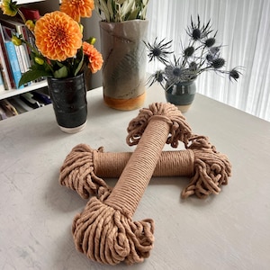 May include: Two spools of thick, natural brown cotton rope, lying on a white surface. The rope is made of multiple strands twisted together, creating a textured look.