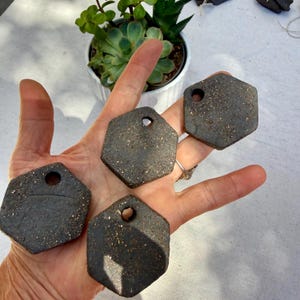 May include: Four dark gray hexagonal ceramic tags with a speckled, textured finish. Each tag features a single circular hole near the top edge, suitable for hanging. The surfaces have a rough, granular appearance with small gold-toned flecks.