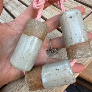 May include: Three ceramic cylindrical objects with a speckled, off-white glaze and a brown, textured base. Each is attached to a pink rope. The objects are held in a hand, with a wooden surface in the background.