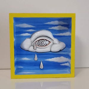 May include: A white cloud with a swirling eye and three teardrops painted on a blue background. The cloud is framed in a yellow wooden frame.