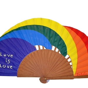 May include: A rainbow-colored hand fan with a wooden frame. The fan is open and the colors are red, orange, yellow, green, blue, and purple. The words "Love is Love" are written on the fan in white. The fan is made by De Arte.