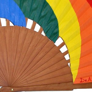 May include: A wooden hand fan with a rainbow design on the fabric. The fan is closed and the fabric is folded in a rainbow pattern. The colors are purple, blue, green, yellow, orange, and red. The fan is signed "DeAnita".