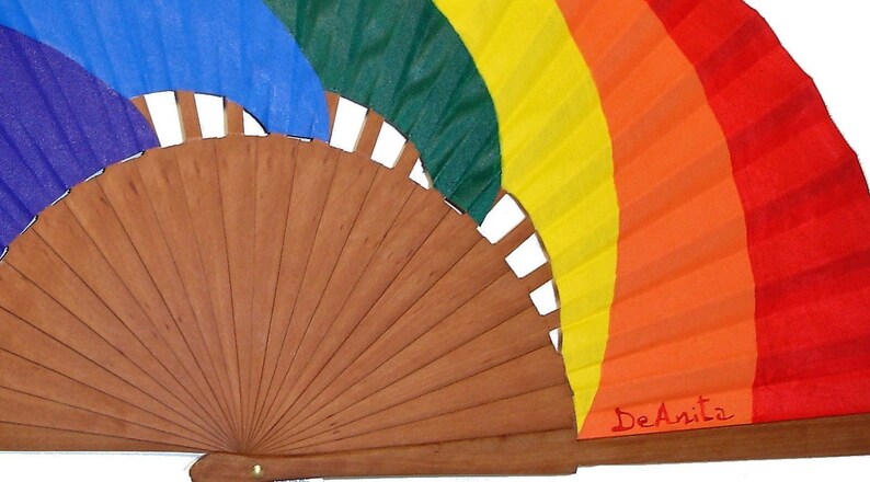 May include: A wooden hand fan with a rainbow design on the fabric. The fan is closed and the fabric is folded in a rainbow pattern. The colors are purple, blue, green, yellow, orange, and red. The fan is signed "DeAnita".