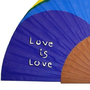 May include: A purple hand fan with a wooden handle. The fan is decorated with a rainbow design and the words "Love is Love" written in white.
