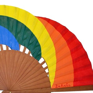 May include: A rainbow-colored hand fan with a wooden frame. The fan is painted with the colors of the rainbow, starting with red at the bottom and ending with yellow at the top. The fan is signed "DeAnita" in red ink.