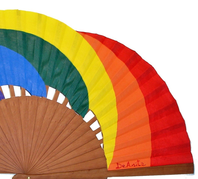 May include: A rainbow-colored hand fan with a wooden frame. The fan is painted with the colors of the rainbow, starting with red at the bottom and ending with yellow at the top. The fan is signed "DeAnita" in red ink.