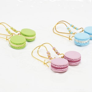 Könnte beinhalten: Drei Paar Hängeohrringe mit Macarons in Pastellfarben. Die Ohrringe sind aus goldfarbenem Metall und haben kleine Perlen am Draht. Die Macarons sind in Grün-, Blau- und Rosatönen.
