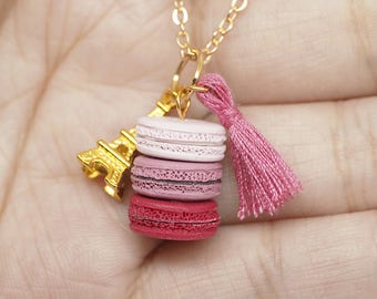 May include: A gold-colored necklace with a charm featuring a stack of pink macarons, a gold Eiffel Tower charm, and a pink tassel. The macarons are in shades of pink and red. The necklace is on a gold chain.