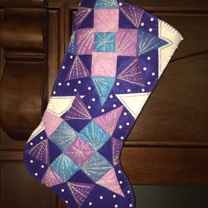 May include: A purple Christmas stocking with a patchwork design of pink, blue, and white triangles and squares. The stocking is decorated with white dots and silver glitter.