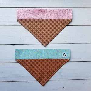 May include: Two dog bandanas with an ice cream cone design. The top bandana is pink with white sprinkles and the bottom bandana is blue with white sprinkles. Both bandanas have a brown waffle cone pattern on the bottom half. The bottom bandana has a white tag with the letter "S" on it.
