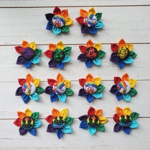 May include: Twelve rainbow-colored felt flower pins with various Disney character designs. The flowers are arranged in three rows of four. The characters include Mickey Mouse, Minnie Mouse, Pluto, Goofy, Donald Duck, and a few others.
