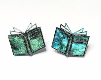 May include: A pair of book-shaped earrings with a teal and green glitter finish. The earrings have a black outline and are open book style.