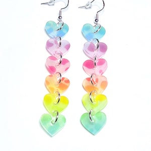May include: Rainbow heart dangle earrings with a silver hook. The hearts are translucent and have a gradient of colours from blue to green to yellow to orange to pink.