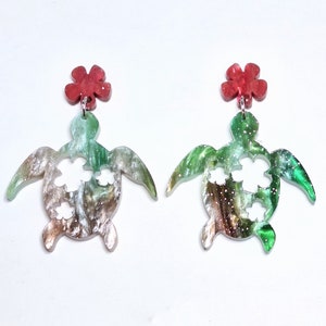 May include: A pair of green and brown turtle earrings with red flower accents. The turtles have cut-out flower designs.
