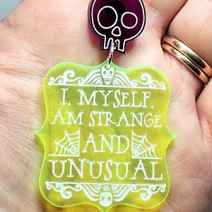 May include: A yellow acrylic charm with the text "I. MYSELF. AM STRANGE AND UNUSUAL" in white lettering. A black skull charm hangs from the top of the charm.