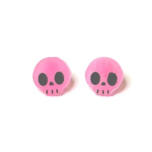 May include: A pair of pink skull-shaped stud earrings. Each earring features a pink skull with black eye sockets and a black nose and mouth. The earrings are set against a white background.