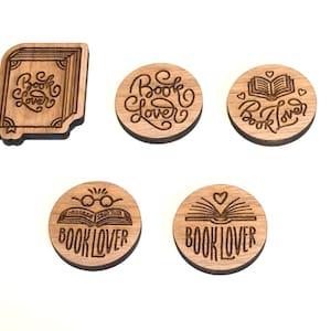Könnte beinhalten: Holzaccessoires zum Thema Bücher mit der Aufschrift "Book Lover". Die Kollektion umfasst einen buchförmigen Lesezeichen und runde Untersetzer mit Buch- und Herzmotiven. Die Artikel sind aus hellbraunem Holz gefertigt.